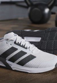 Λευκό και μαύρο αθλητικό παπούτσι Adidas με τρεις μαύρες ρίγες, που βρίσκεται σε ξύλινο πάτωμα με εξοπλισμό γυμναστηρίου θολωμένο στο φόντο.