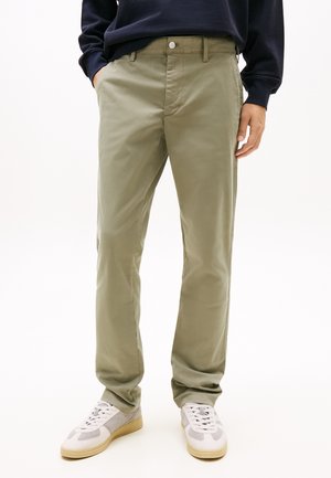 Muškarac u bež hlačama ravnog kroja, bijelim i sivim sportskim cipelama te tamnoj majici s dugim rukavima, s rukama u džepovima.