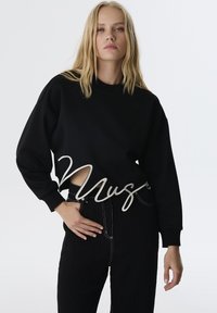 Suéter negro recortado con mangas oversize que presenta un diseño de corte y letras bordadas en blanco. Combinado con pantalones negros de cintura alta.