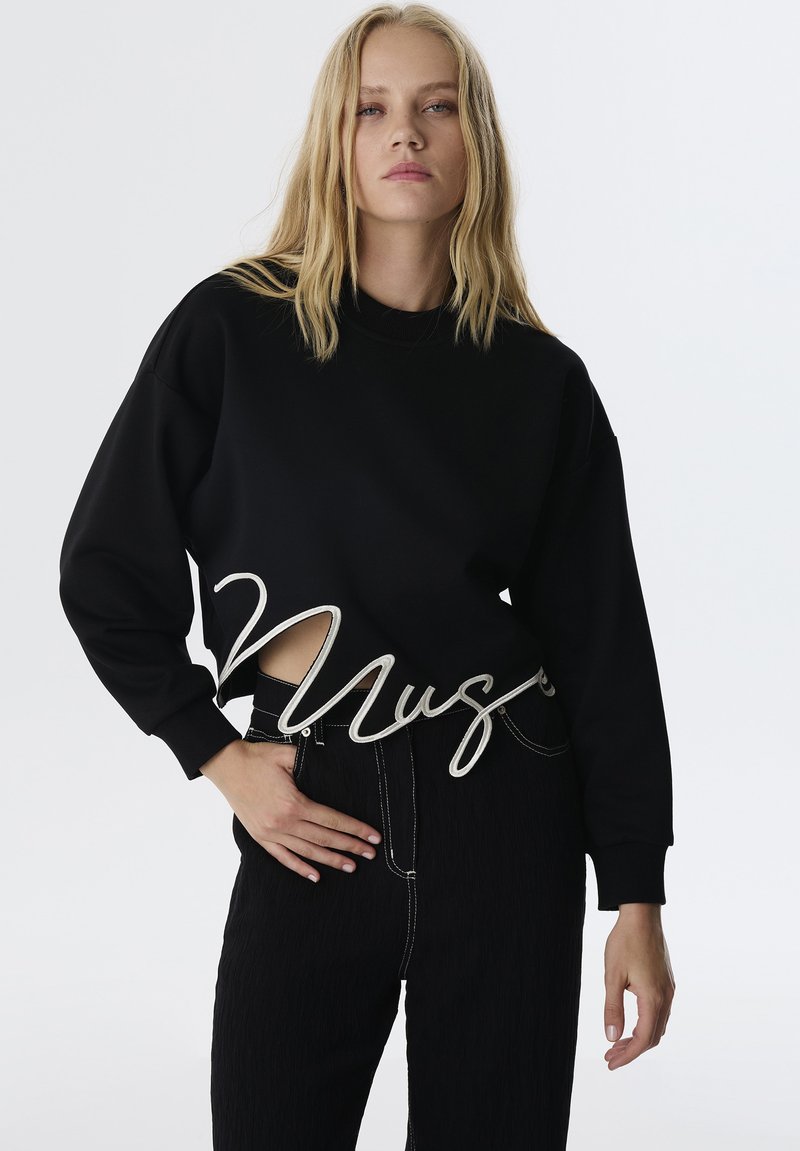 Suéter negro recortado con mangas oversize que presenta un diseño de corte y letras bordadas en blanco. Combinado con pantalones negros de cintura alta.