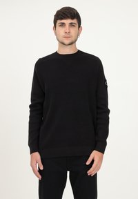 Maglione nero lavorato a maglia con colletto rotondo e maniche lunghe. Presenta un motivo testurizzato e una piccola etichetta con logo sulla manica sinistra.