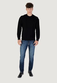 Maglione nero a maglia con motivo testurizzato, scollatura rotonda, polsini a coste, abbinato a jeans blu e sneakers nere, in piedi davanti a uno sfondo neutro.