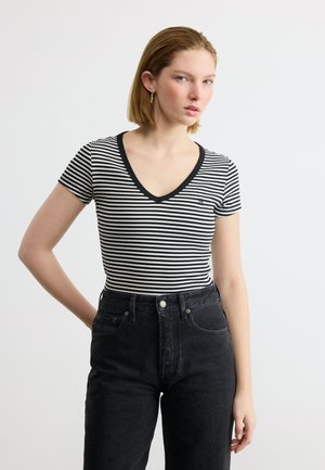 Ung kvinde med kort blondt hår iført sort-hvid stribet V-hals skjorte og højtaljede sorte jeans mod en ensfarvet baggrund.