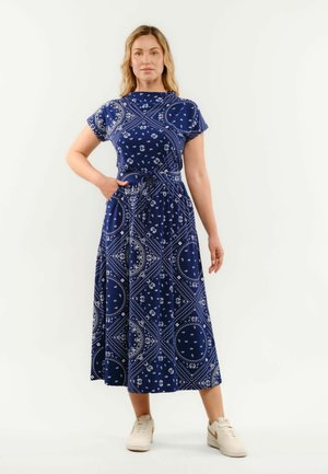 Žena v modrom vzorovanom midi šatách s krátkymi rukávmi a bielych teniskách, stojaca s jednou rukou v vrecku na bielom pozadí.