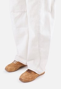 Chaussures en daim fauve avec une surface texturée et des accents lisses. Associées à un pantalon blanc ample en coton avec des détails de couture visibles.