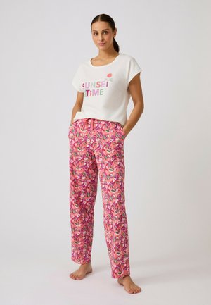 Femme aux cheveux foncés en queue de cheval portant un T-shirt blanc avec le texte "SUNSET TIME" et un pantalon rose à motif floral coloré, debout pieds nus.