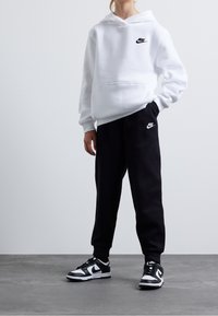 Witte Nike hoodie met voorzak en logo, gedragen met zwarte Nike joggers met logo. Schoenen: zwart-witte Nike sneakers.
