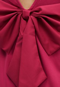 Suur punane kangasniit kaelaaugu juures, seotud punase riideeseme keskpunktis rinnal, nähtavate kangasvoltide ja õmblustega.