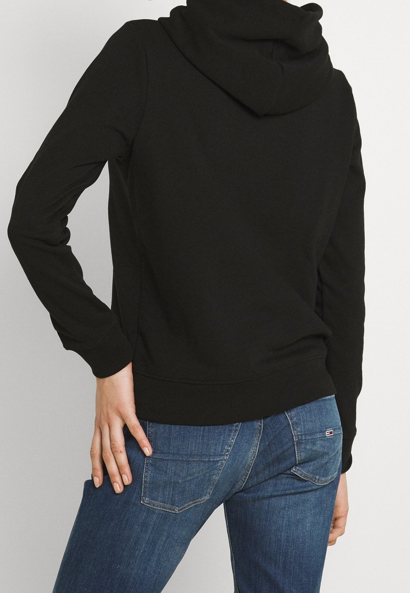 Osoba v černé mikině s kapucí a modrých džínách, viděná ze zadu, s jednou rukou položenou na boku.