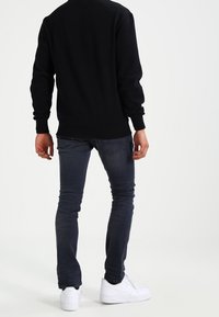 Sweat-shirt noir à manches longues, associé à un jean en denim foncé et des baskets blanches. Tissu lisse, coupe ajustée, détails de couture visibles.