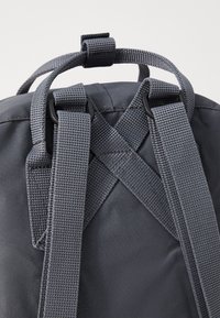 Sivi ruksak s debelim, teksturiranim trakama koje se preklapaju na leđima. Čini se da je materijal izdržljiv, s glatkom površinom i čvrstim detaljima od hardvera.