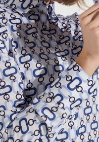 Košile se zapínáním v bílé látce, zdobená geometrickým řetězcem v modrých a hnědých tónech. Hladká textura s klasickým límečkem a manžetami.