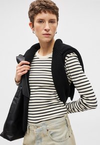 Μαύρο πλεκτό γιλέκο πάνω από ένα μπεζ και μαύρο ριγέ πουκάμισο με μακριά μανίκια. Ανοιχτό μπλε τζιν παντελόνι, κρατώντας μια μαύρη δερμάτινη τσάντα.