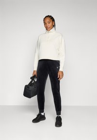 Witte cropped sweatshirt met een hoge kraag, gecombineerd met marineblauwe fluwelen joggers en zwarte sneakers. Een zwarte sporttas wordt in één hand vastgehouden.