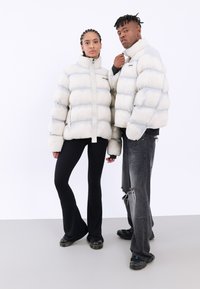 Två individer som bär stora, puffiga, ljusgrå jackor med dragkedja framtill och svarta accenter. Den ena har på sig svarta utsvängda byxor, den andra slitna jeans.