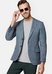 Niebieski marynarka w kratę z dwoma guzikami, z klapami i dwiema kieszeniami, noszona na szarym t-shircie i ciemnozielonych spodniach.