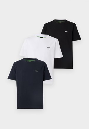 Jeune homme debout, main dans la poche, portant un T-shirt noir BOSS, un jean bleu clair et des baskets blanches sur fond gris uni.