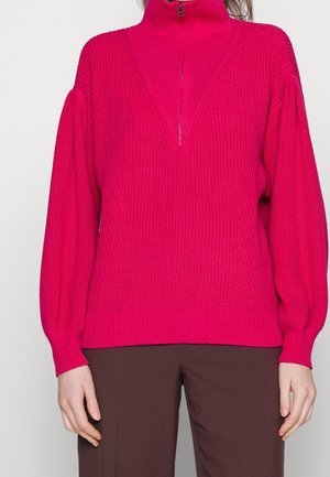 Pull en maille rose vif avec col montant à zip et manches longues bouffantes porté avec un pantalon marron foncé sur une personne.