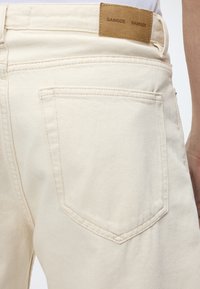 Κοντινή λήψη πίσω τσέπης από εκρού τζιν παντελόνι με ταμπέλα από γνήσιο δέρμα χρώματος ταμπά με την επιγραφή "SAMSOE SAMSOE" στη μέση.