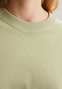 T-shirt di cotone verde chiaro con colletto a costine e frase ricamata "FAI PARTE DELLA MIA STORIA" in bianco. Texture liscia.