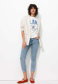 Blazer blanc, t-shirt graphique avec du texte bleu, jeans bleu clair et ballerines rouges. Tissus doux, coupe ajustée, manches retroussées et semelles plates.