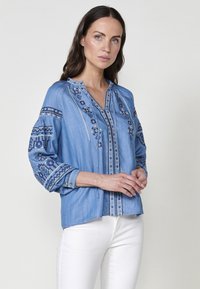 Ljusblå denimblus med intrikata blå broderier. Har V-ringning, lång ärm med accentmönster och en avslappnad passform.