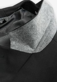 Schwarzer Kapuzenpullover mit einem grauen Filzkraus. Das Innere zeigt ein gemustertes Futter. Detaillierte Nähte am Kragen und Halsausschnitt.