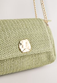 Sac à main tissé vert clair avec une fermeture ronde irrégulière dorée et une bandoulière chaîne dorée sur un fond neutre.