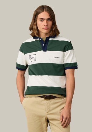 Jonge man met schouderlang haar, die een groen-wit gestreept poloshirt en beige broek draagt tegen een lichtgrijze achtergrond.