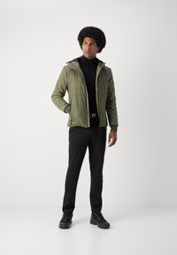 Veste légère olive avec texture matelassée et capuche ajustable, associée à un pantalon noir et des baskets noirs.