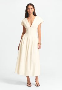 Kermansövuinen puuvillamekko, jossa on syvä V-pääntie, lyhyet hihattomat hihat ja pitkään valuva pituus. Mekossa on istuva yläosa ja laskostettu hame.