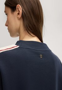 Navyblauwe polo met een textuurstof, voorzien van een henley-kraag, rode en crèmekleurige strepen op de schouders en een klein logo bij de hals.
