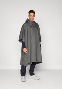 Poncho gris imperméable avec capuche, présentant une coupe ample, des ouvertures latérales et un design minimaliste. Porté par-dessus un pantalon et des chaussures noirs.