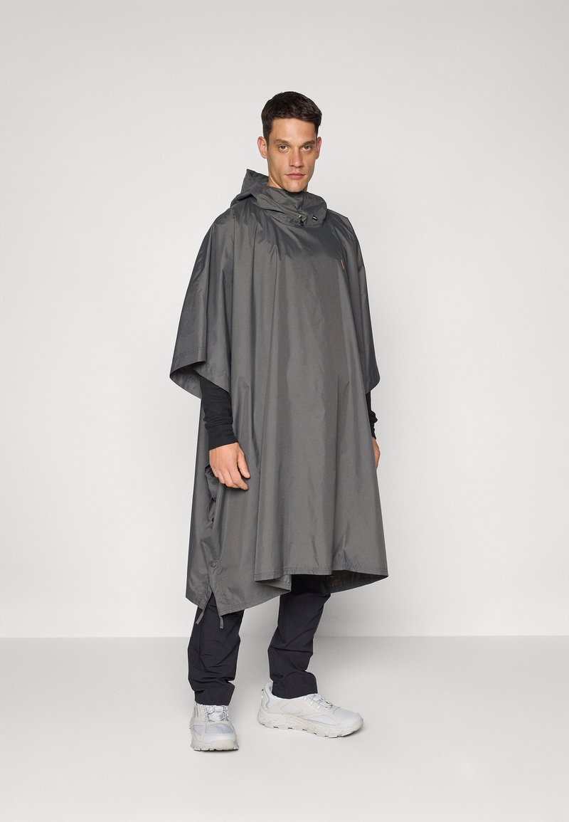 Poncho gris imperméable avec capuche, présentant une coupe ample, des ouvertures latérales et un design minimaliste. Porté par-dessus un pantalon et des chaussures noirs.