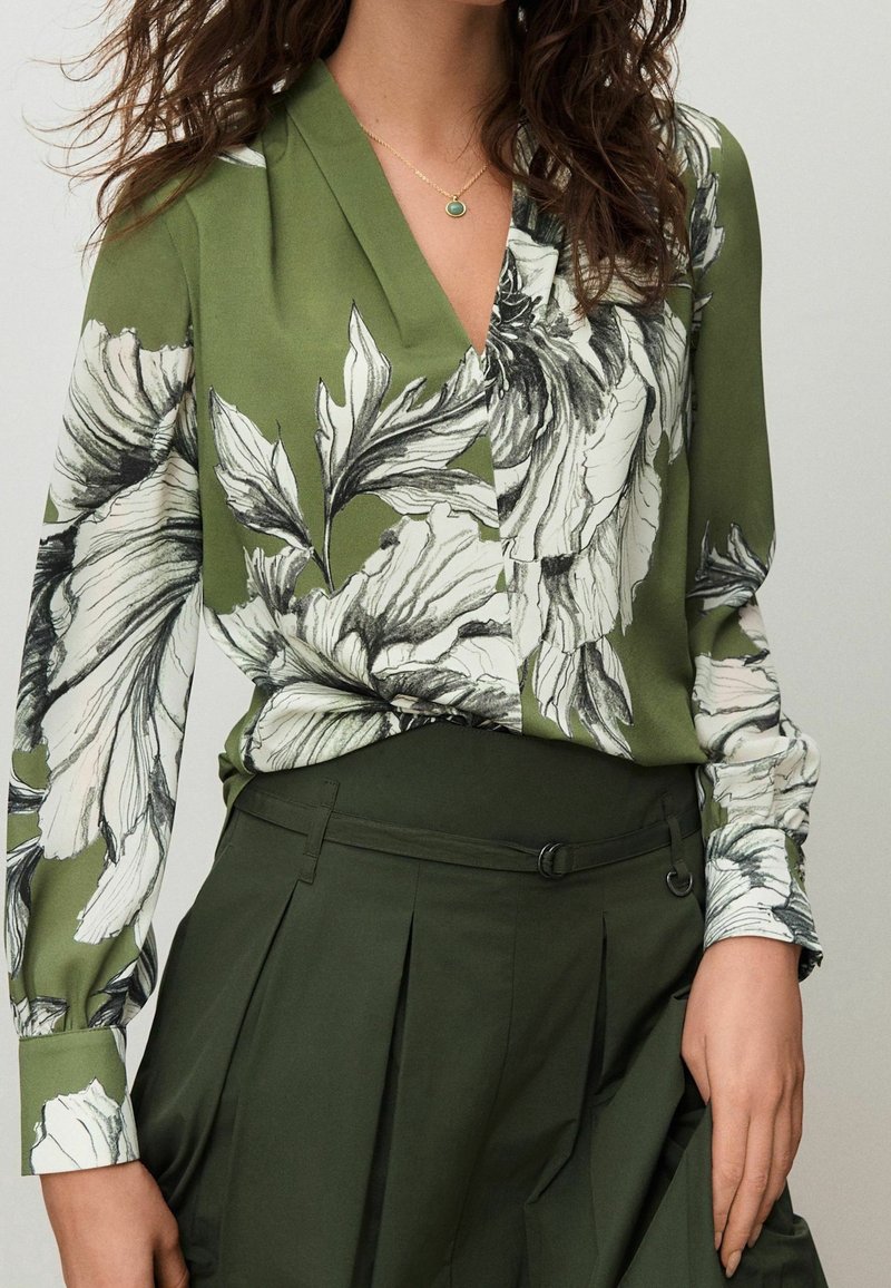 Femme portant un chemisier vert olive avec de grands imprimés floraux blancs et noirs et un pantalon plissé taille haute vert foncé.