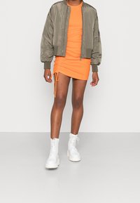 Giacca bomber verde oliva, mini abito arancione con arricciature laterali e stivali da combattimento bianchi con lacci e suole spesse.