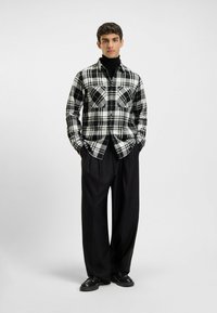 Zwart-wit geruite flanellen overhemd met twee borstzakken, gedragen met losse zwarte broek en zwarte schoenen. Glad textuur, relaxte pasvorm.