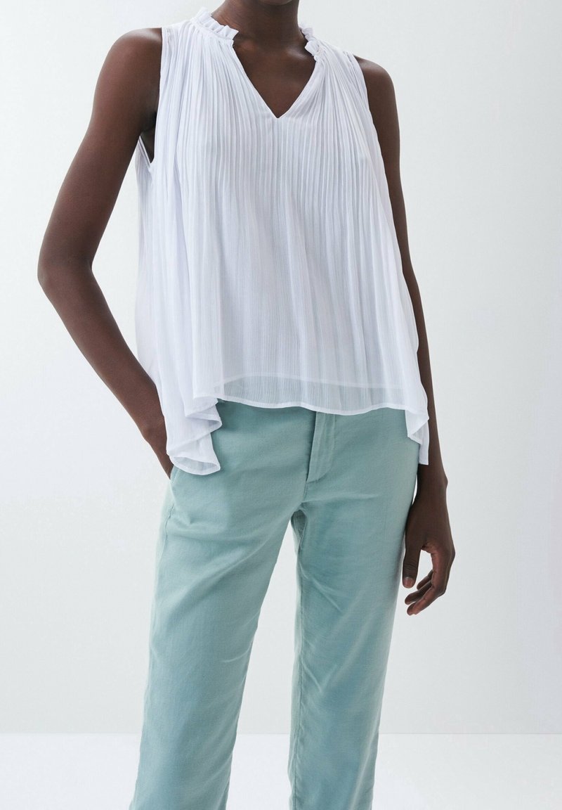 Personne à la peau foncée portant un chemisier blanc plissé sans manches et un pantalon bleu clair, main reposant dans la poche sur fond uni.