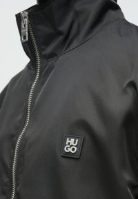 Μαύρο μπουφάν με φερμουάρ και ανεβασμένο ασημένιο λογότυπο "HUGO" στο στήθος, με ψηλό γιακά και λεπτομερή έλκτη φερμουάρ.