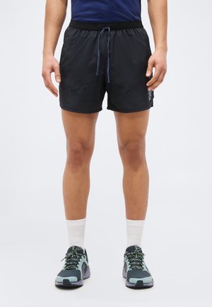 Άνδρας που φοράει μαύρο αθλητικό σορτς, λευκές κάλτσες και γκρι-πράσινα παπούτσια τρεξίματος, στέκεται μπροστά σε απλό φωτεινό φόντο.