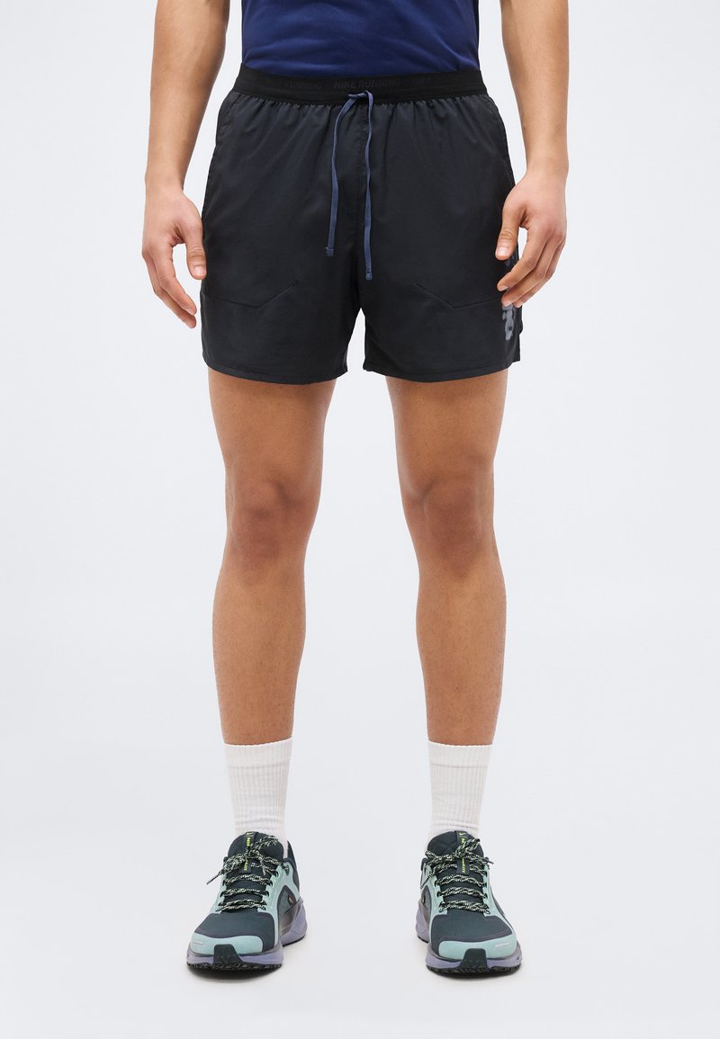 Άνδρας που φοράει μαύρο αθλητικό σορτς, λευκές κάλτσες και γκρι-πράσινα παπούτσια τρεξίματος, στέκεται μπροστά σε απλό φωτεινό φόντο.