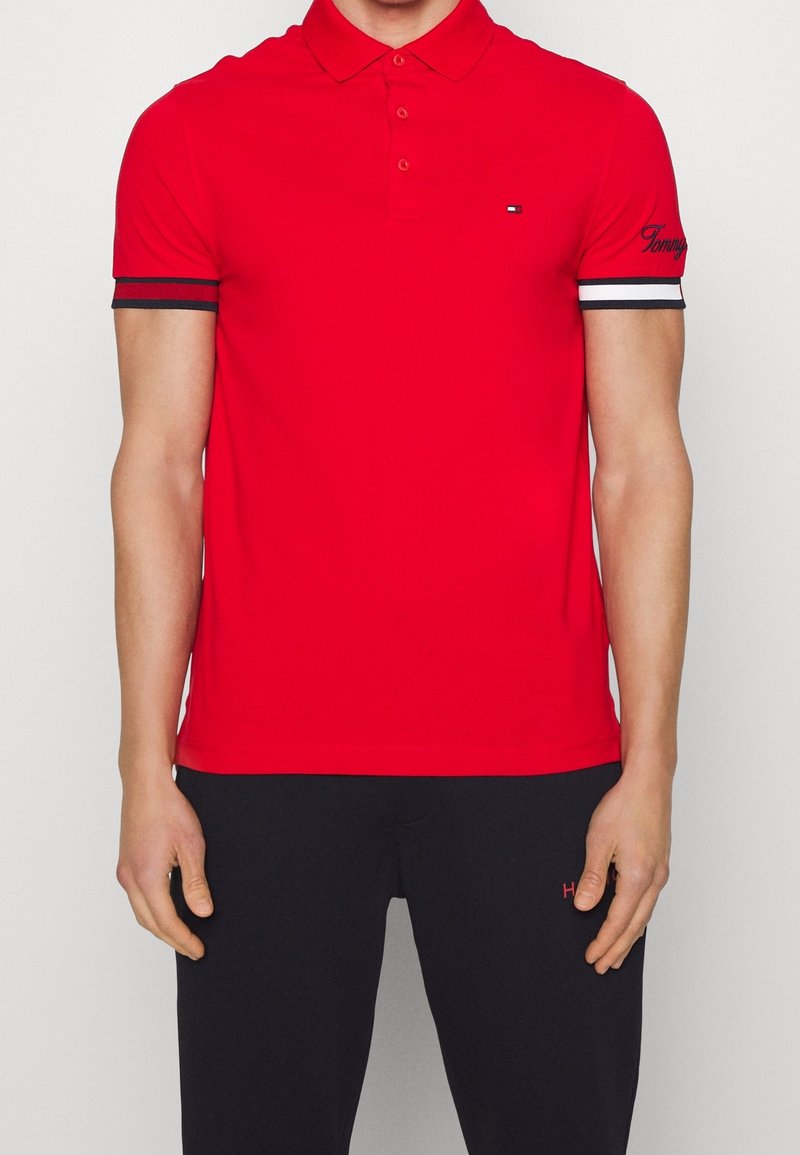 Homme portant un polo rouge avec des poignets de manches rayés noir, blanc et rouge, et un pantalon noir, debout les bras détendus.
