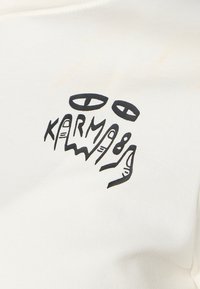 Weißes Baumwoll-T-Shirt mit einem schwarzen Grafikdesign von Händen und Augen, in das der Text "KARM" integriert ist. Glatte Textur, lässiger Schnitt.