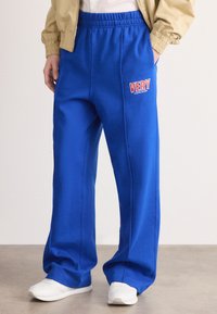 Persona, kas tērpta brīvās zils biksēs ar uzrakstu "VERY Calvin Klein" uz augšstilba, baltās sporta apavās, bēšā jakā un baltā krekla, stāv uz betona grīdas.