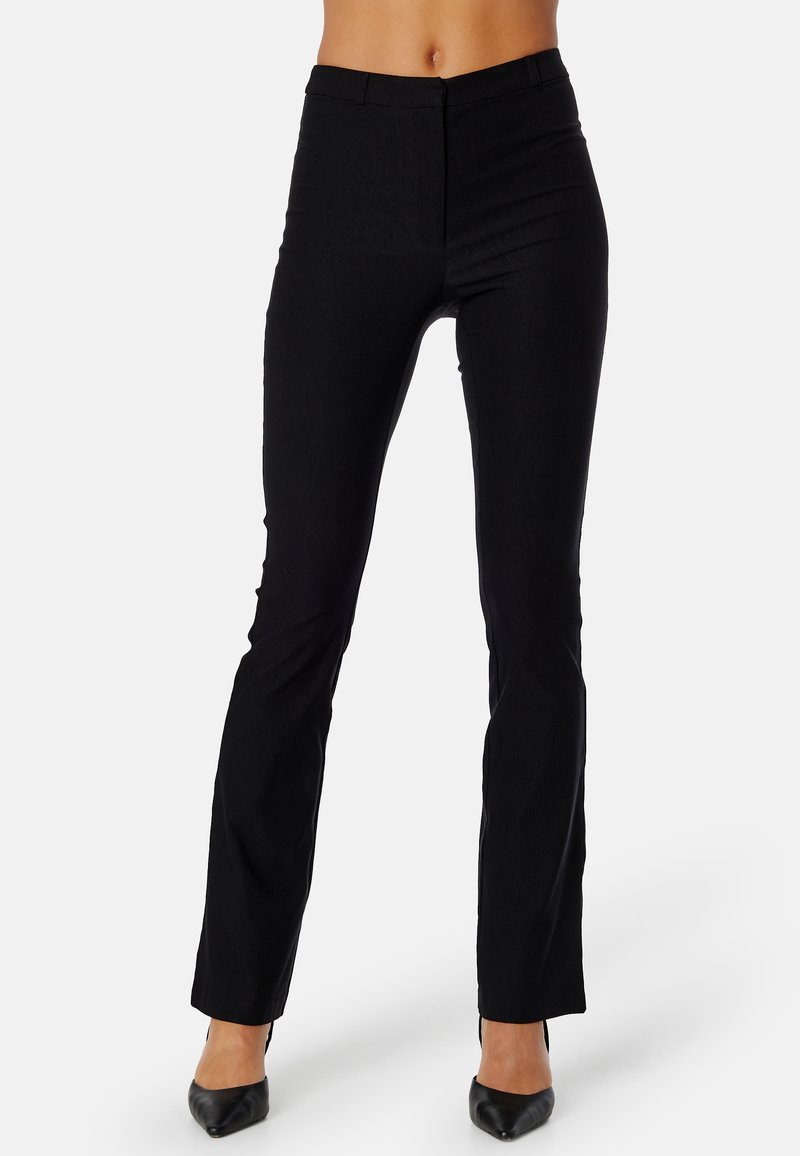 Femme portant un pantalon noir ajusté et des talons noirs à bouts pointus, debout devant un fond clair uni.