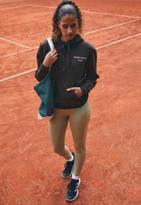 Rudas kapučinas su logotipu, šviesiai rudi leggingai ir juodi sportbačiai. Laiko melsvą krepšį. Fone yra raudonos molio teniso aikštelės.