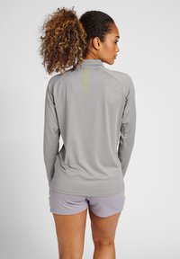 Maglia atletica grigia a maniche lunghe con colletto alto, caratterizzata da un dettaglio del logo riflettente. Il tessuto è liscio, con un design aderente e maniche raglan.