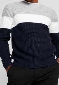 Knitted crewneck sweater featuring horizontal stripes in grey, white, and navy. Textured fabric with ribbed cuffs and hem.