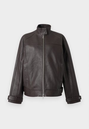 Žena so širokou tmavo hnedou koženou bundou, bielou košeľou, tmavými širokými džínsami, čiernymi špicatými podpätkami, slnečnými okuliarmi a náušnicami v tvare kruhu.