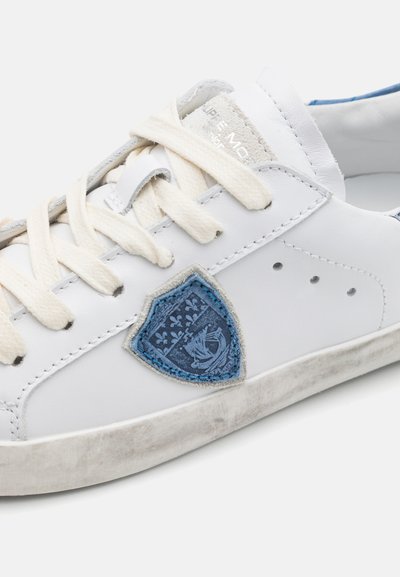 Weißes Leder-Sneaker mit beigen Schnürsenkeln, versehen mit einem blauen, gestickten Wappen in Schildform und hellgrauen Akzenten an Ferse und Sohle.